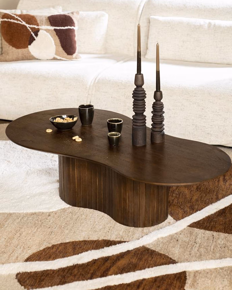 Een donkerhouten organische tafel met een organische, ronde vorm staat op een beige gedessineerd tapijt. Op de tafel staan twee hoge decoratieve kandelaars, twee kleine kopjes en een schaal met hapjes. Een witte bank staat op de achtergrond.