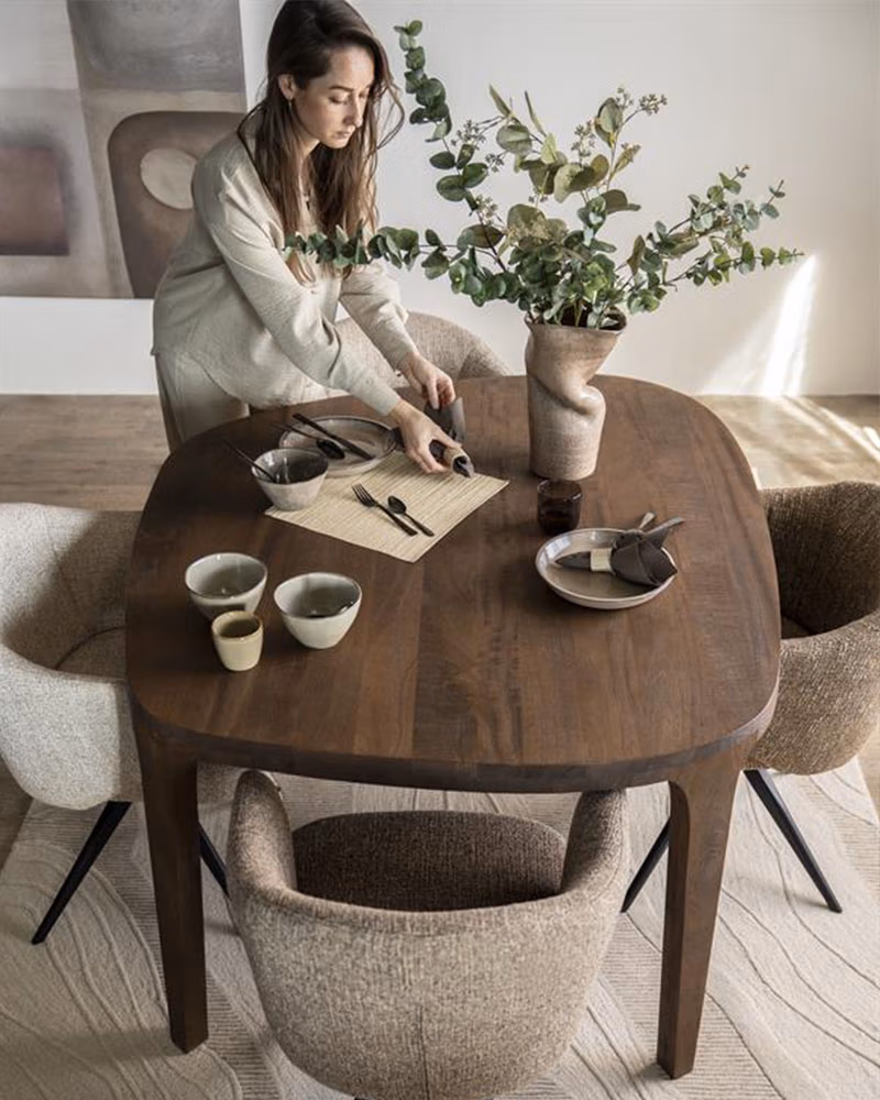 Een vrouw zet een organische tafel neer, versierd met een vaas met groen, kommen, borden en bestek, omringd door vier gestoffeerde stoelen in een moderne, gezellige eetkamer.