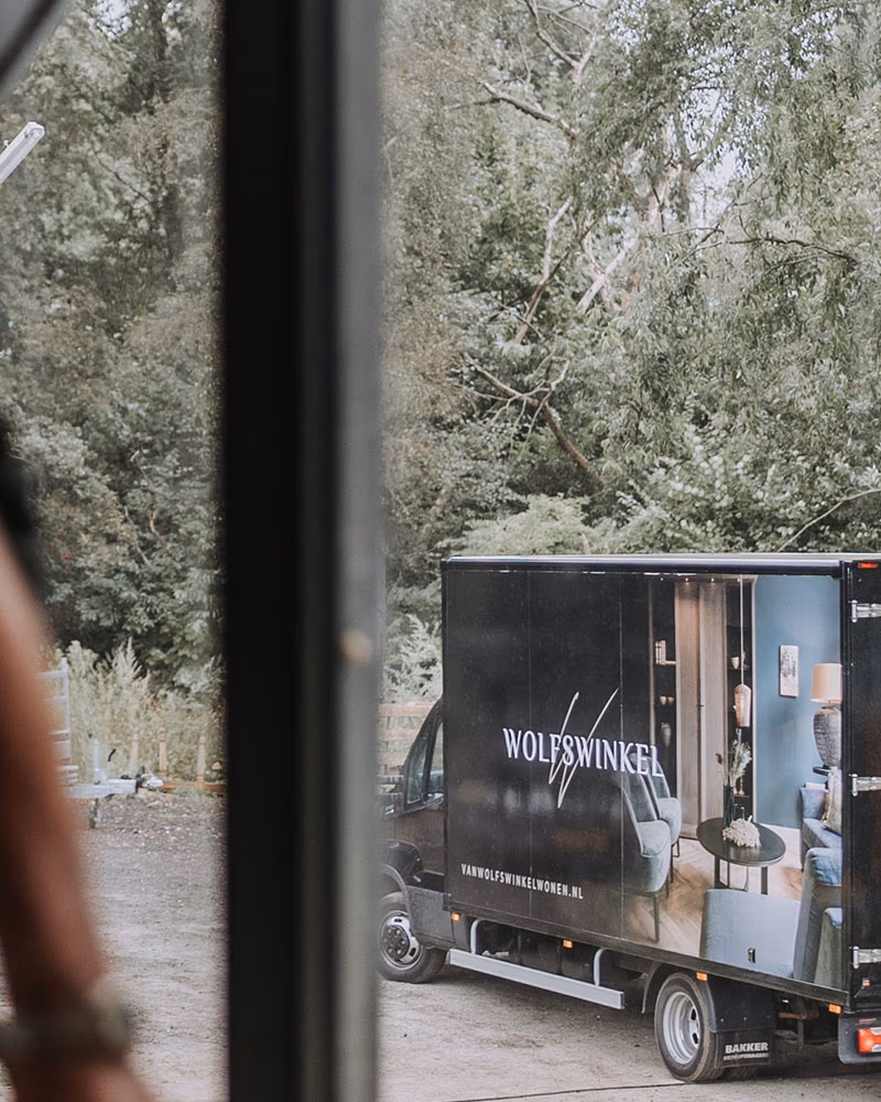 Een zwarte Wolfswinkel bestelwagen met reclame voor woninginrichting en organische tafel staat geparkeerd op een grindvlakte bij bomen, gezien door een raamkozijn vanuit een gebouw.
