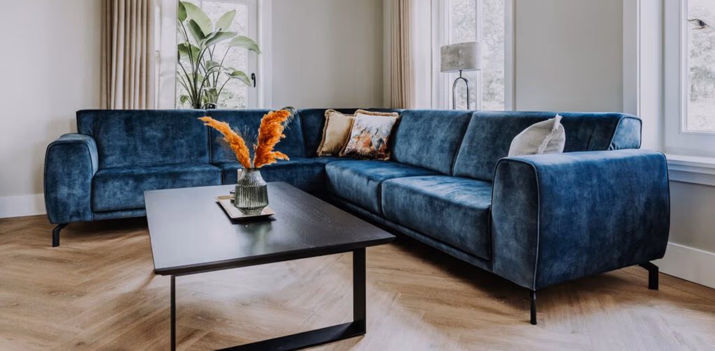 Een moderne woonkamer met een blauwe fluwelen hoekbank, een rechthoekige, zwarte salontafel met een oranje bloemstuk, een hoge groene plant, een lichte houten visgraatvloer en grote ramen met beige gordijnen.
