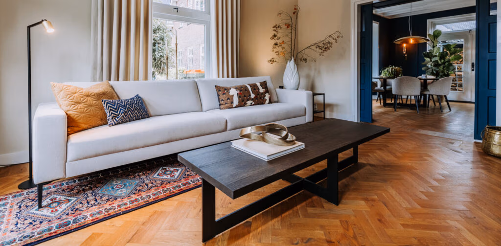 Moderne woonkamer met een witte bank, kussens met patroon, een staande lamp, een houten salontafel met boeken en decoraties, een kleed en uitzicht op de eethoek met een tafel, stoelen en planten.