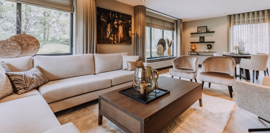 Moderne woonkamer met beige banken, fauteuils, een donkere houten salontafel, grote ramen met gordijnen, wanddecoratie en decoratieve objecten. Zachte neutrale tinten en overvloedig natuurlijk licht creëren een gezellige, elegante sfeer.