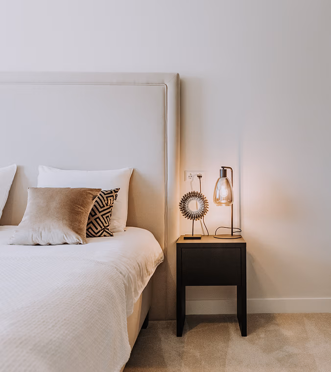 Een moderne slaapkamer met een netjes opgemaakt bed, beige hoofdeinde, decoratieve kussens en een zwart nachtkastje met een lamp en een kleine decoratieve spiegel tegen een eenvoudige witte muur.