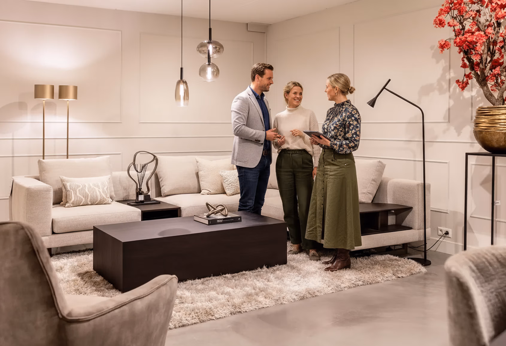 Drie mensen staan te praten in een moderne, stijlvolle woonkamer met neutraal gekleurd meubilair, een pluche vloerkleed, eigentijdse verlichting en decoratieve accenten. Eén vrouw houdt een tablet vast terwijl de anderen glimlachen en luisteren.