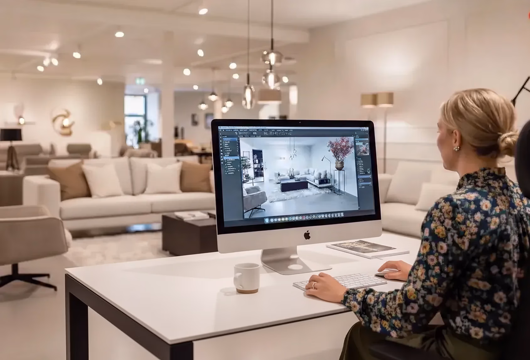 Een vrouw zit aan een bureau in een moderne, goed verlichte meubelshowroom en werkt aan een desktopcomputer waarop een programma voor het bewerken van interieurs wordt weergegeven met een scène in een woonkamer.