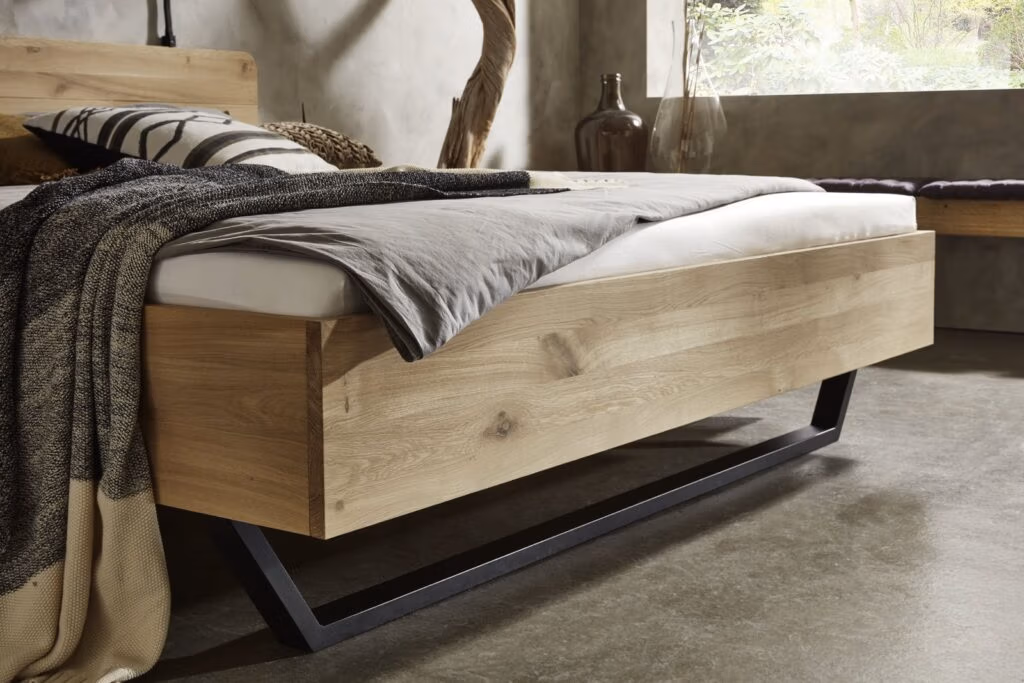 Een modern houten bed met zwarte metalen poten, netjes opgemaakt met grijs en zwart beddengoed, in een minimalistische slaapkamer met betonnen vloeren en een groot raam dat natuurlijk licht binnenlaat.
