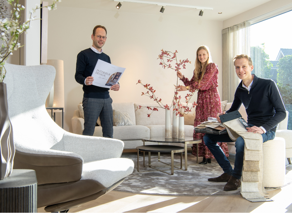 Drie mensen in een lichte, moderne woonkamer. Een man houdt een tijdschrift vast, een vrouw schikt decoratieve takken en een andere man zit met stofstalen. Neutrale tinten en natuurlijk licht vullen de ruimte.