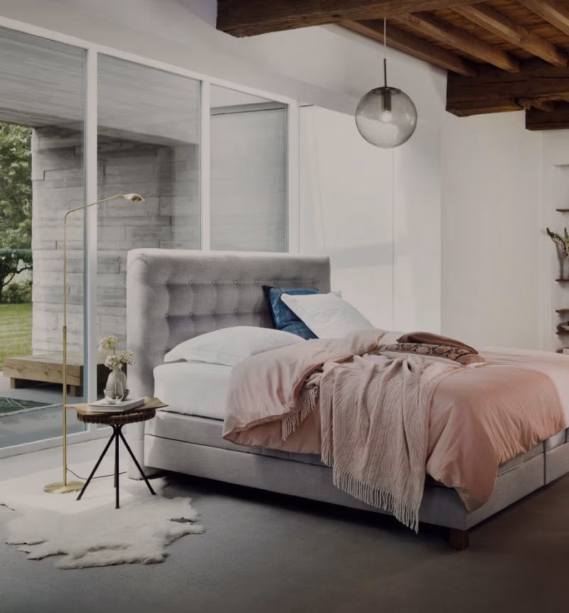 Moderne slaapkamer met een grijs gecapitonneerd bed, lichtroze beddengoed, een rond nachtkastje met een lamp en bloemen, een koeienhuidkleed en grote ramen die uitzicht bieden op een groene buitenruimte.