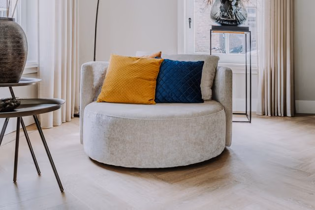 Een ronde, lichtgekleurde fauteuil met een oranje en een blauw kussen staat in een lichte kamer met een houten vloer, bij een raam en neutrale gordijnen. Er staan ook een tafeltje en een plantenstandaard.