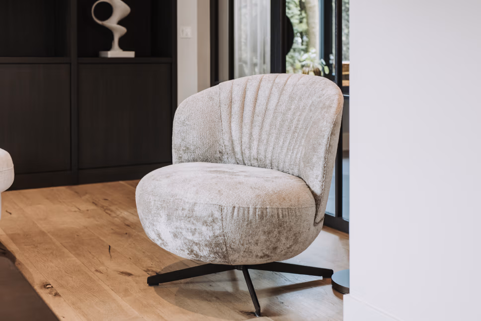 Een moderne, lichtgrijze draaistoel met een gebogen rugleuning staat op een houten vloer, vlakbij grote glazen deuren en donkere kasten in een eigentijdse kamer.
