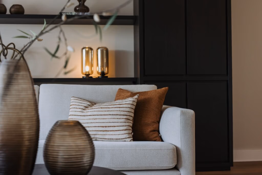 Een moderne woonkamer met een lichtgrijze bank, twee bruine sierkussens, een gestreept kussen, een donkere ronde salontafel met vazen en een plank met twee verlichte glazen lampen op de achtergrond.