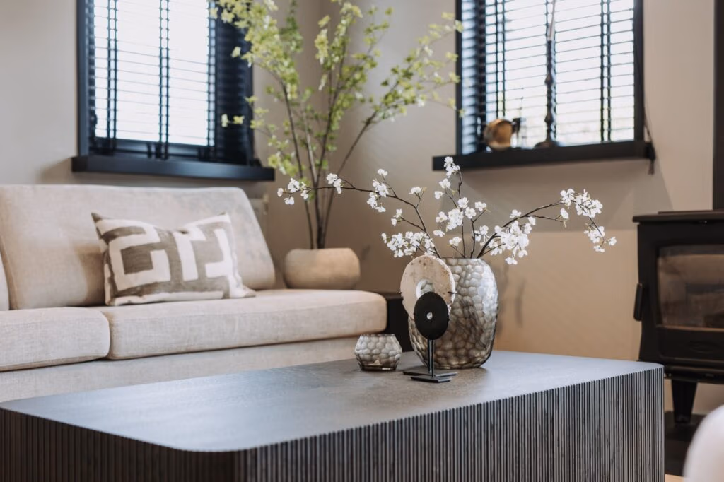 Moderne woonkamer met een lichte bank, geometrisch kussen, zwarte salontafel, vazen met witte bloemen, plant in pot, zwarte kachel en ramen met zwarte jaloezieën die natuurlijk licht binnenlaten.