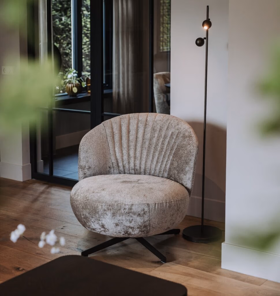 Een gezellige, grijze fauteuil met structuur staat op een houten vloer naast een hoge, slanke zwarte vloerlamp in een moderne woonruimte met grote ramen en groen buiten.