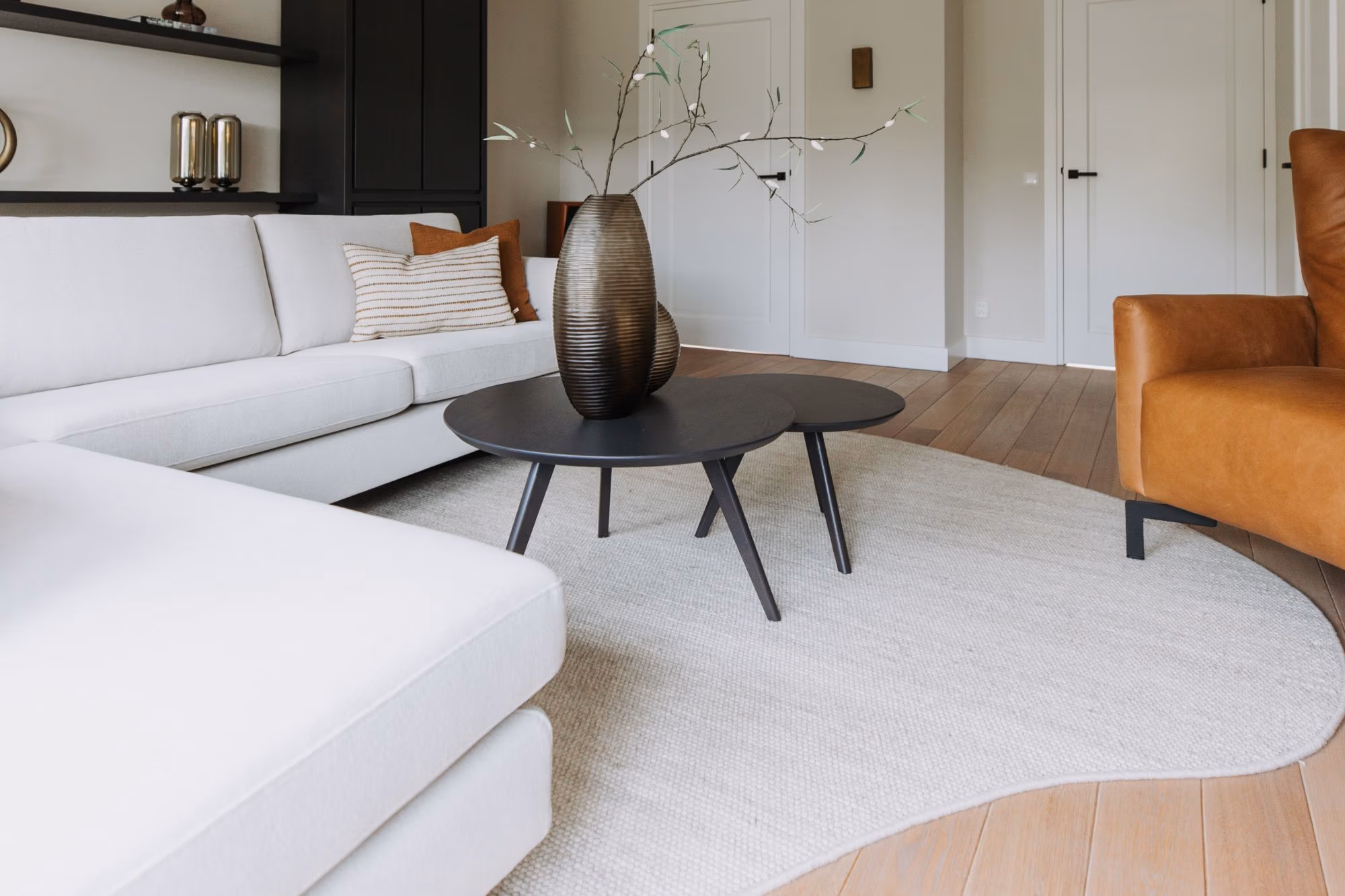 Moderne woonkamer met een witte sectionele bank, bruine leren stoel, ronde zwarte salontafels, een decoratieve vaas met takken en een lichtbeige vloerkleed op een houten vloer. Minimalistische inrichting en neutrale tinten.
