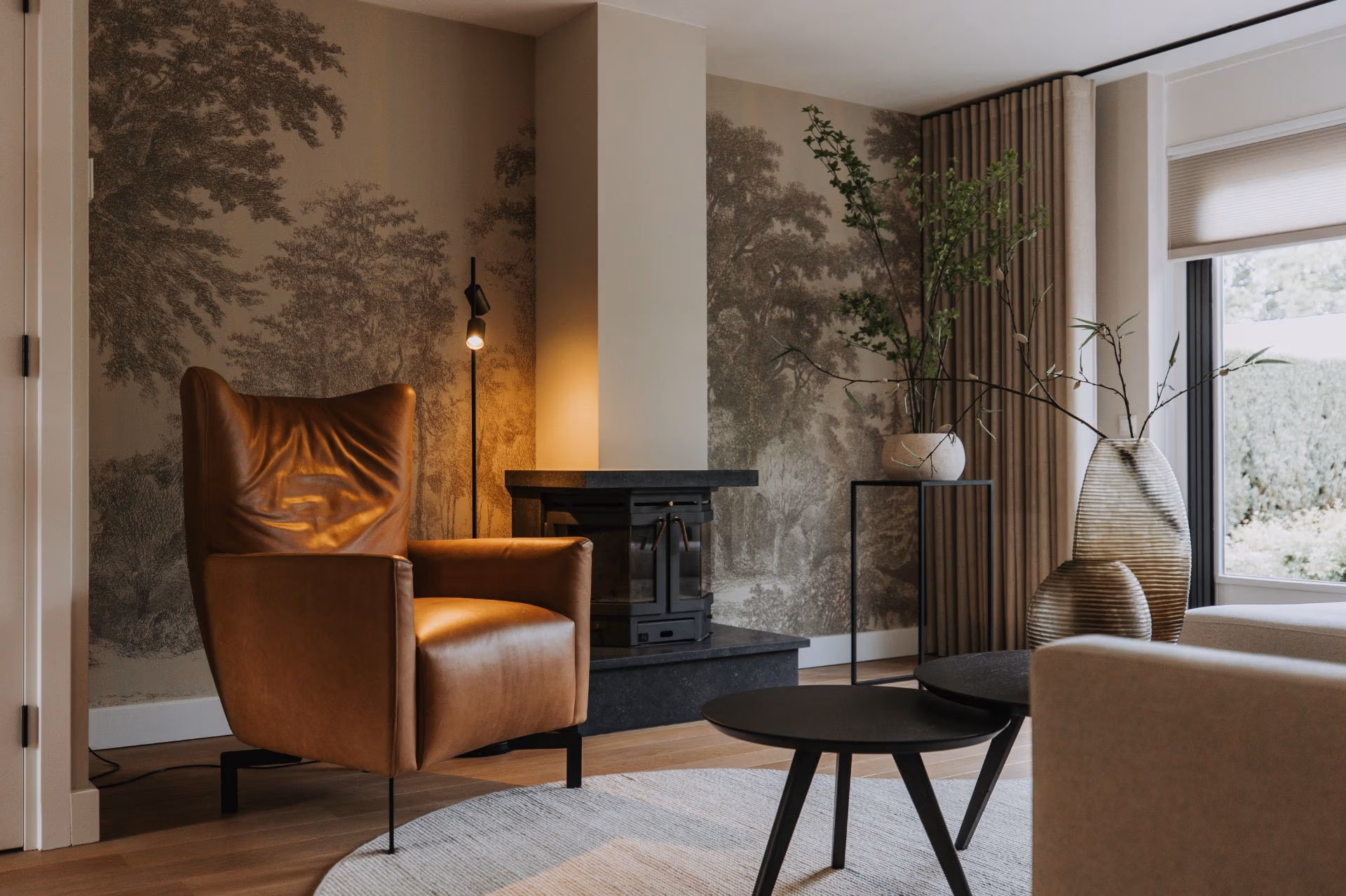 Een moderne woonkamer met een bruine leren fauteuil, zwarte staande lamp, ronde salontafel, decoratieve vazen met takken en een neutrale muurschildering met bomen en natuur. Licht stroomt naar binnen door een groot raam.