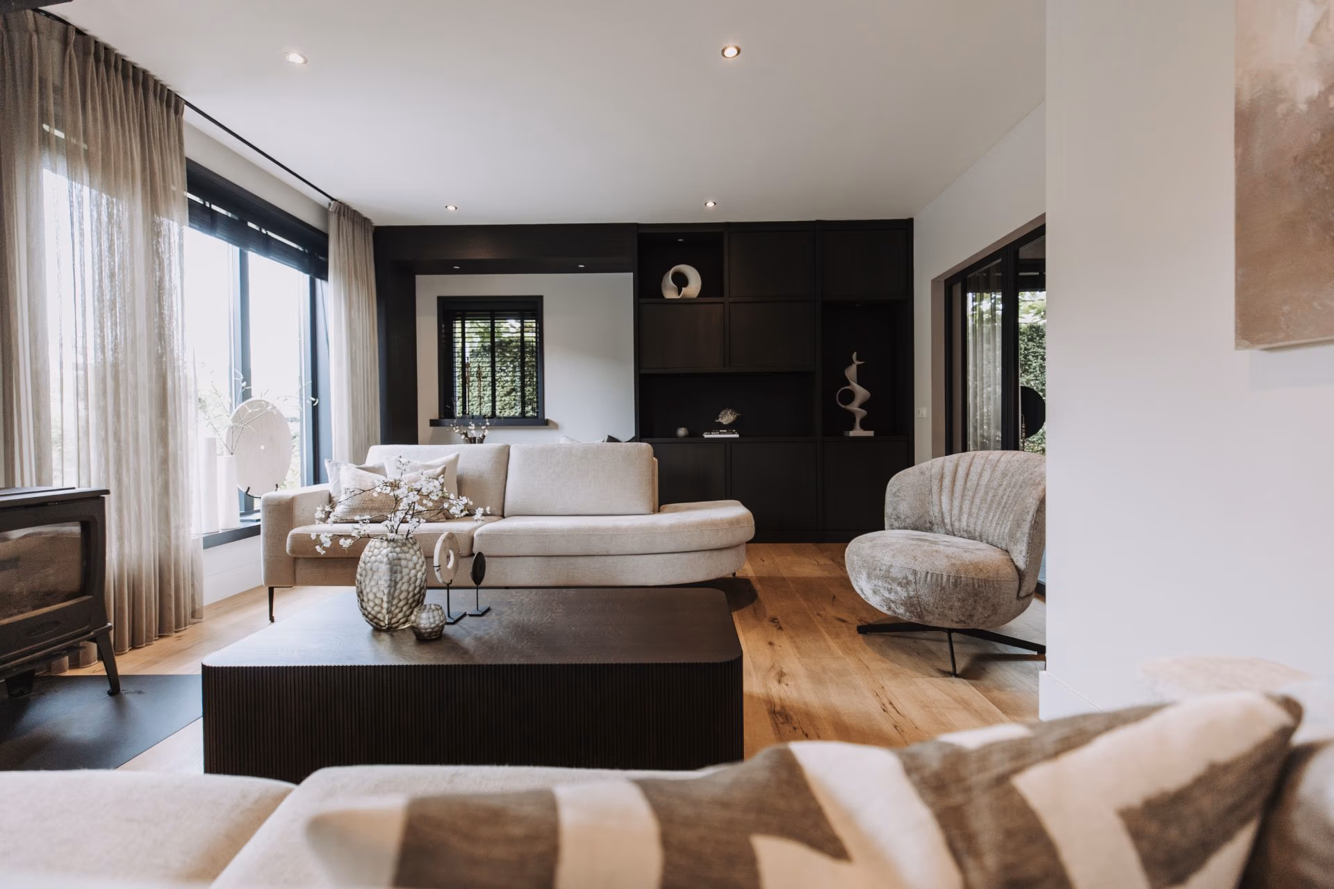 Moderne woonkamer met neutraal getinte meubels, grote ramen met transparante gordijnen, een lichte houten vloer, donkere ingebouwde planken, een salontafel en decoratieve vazen die een gezellige, eigentijdse sfeer creëren.