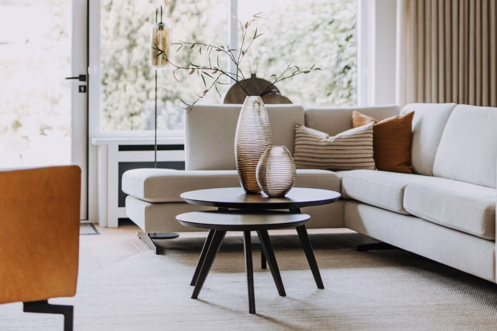 Een moderne woonkamer met een lichtgrijze sofa, neutrale kussens, een ronde zwarte salontafel, decoratieve glazen vazen en grote ramen die natuurlijk licht binnenlaten.