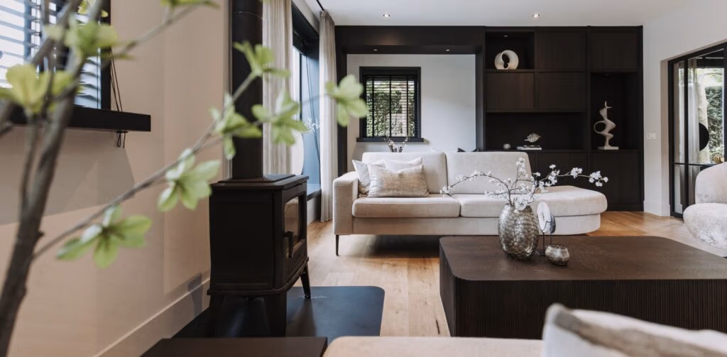 Moderne woonkamer met lichthouten vloer, beige bank, donkerhouten planken, grote ramen en decoratieve vazen met bloemen op een donkere salontafel. Het licht stroomt naar binnen en creëert een heldere en uitnodigende sfeer.