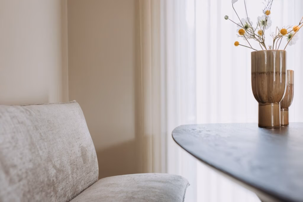 Een beige sofa staat naast een donkere ronde tafel met een bruine keramieken vaas met gedroogde bloemen, tegen dunne witte gordijnen in een zacht verlichte kamer.