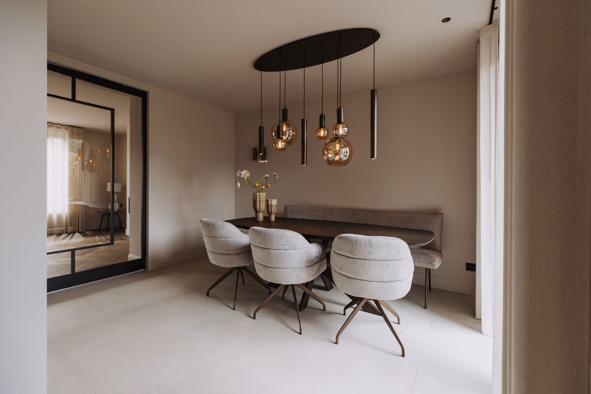 Moderne eetkamer met een donkere ovale tafel, vier gecapitonneerde beige stoelen en verschillende gouden hanglampen die erboven hangen. Grote spiegel aan de linkermuur en transparante gordijnen laten natuurlijk licht van rechts binnen.