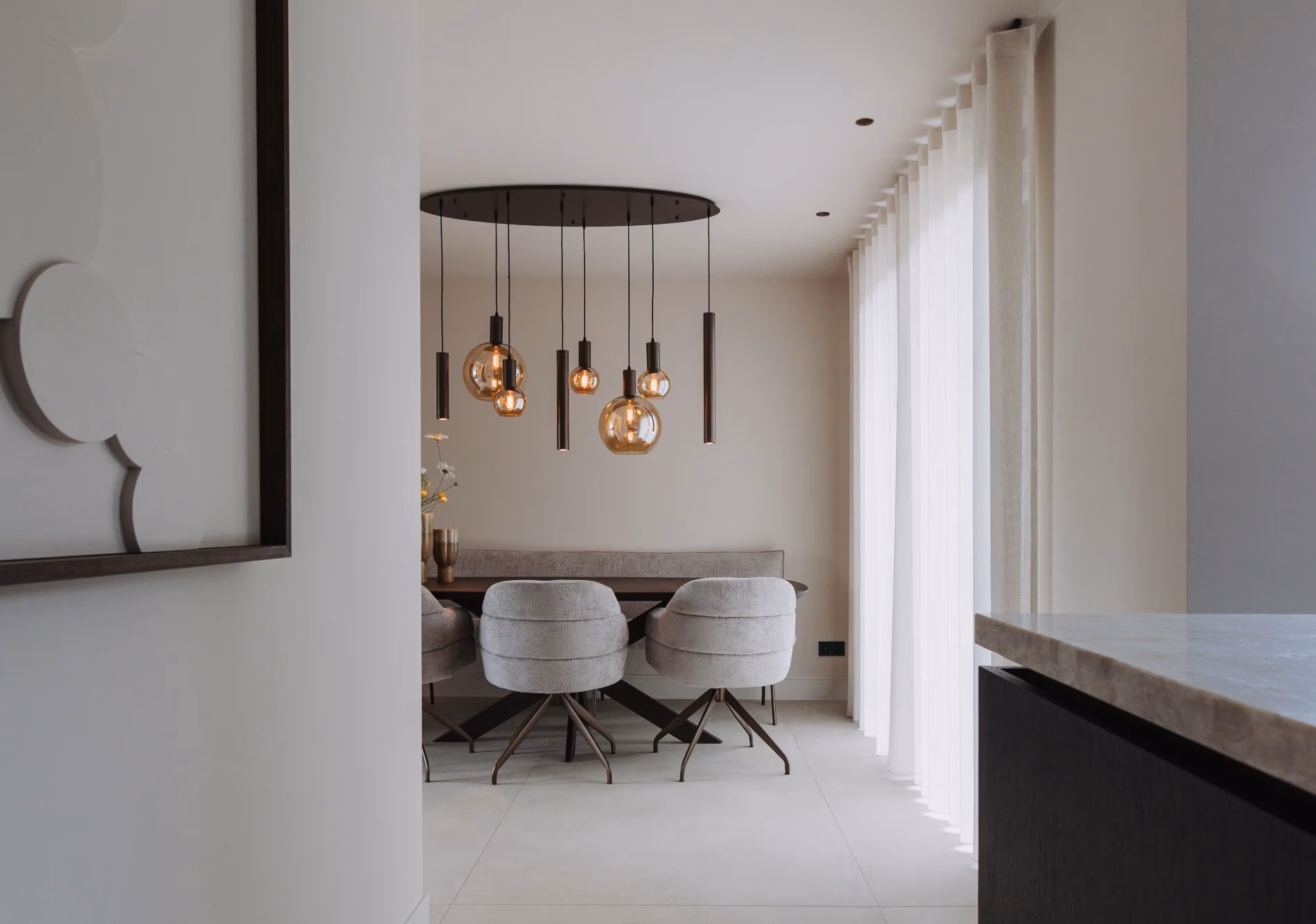 Moderne eethoek met een ronde tafel, vier gestoffeerde stoelen en een set hanglampen met amberkleurige glazen bollen. Grote gordijnen laten natuurlijk licht binnen en creëren een heldere, minimalistische sfeer.