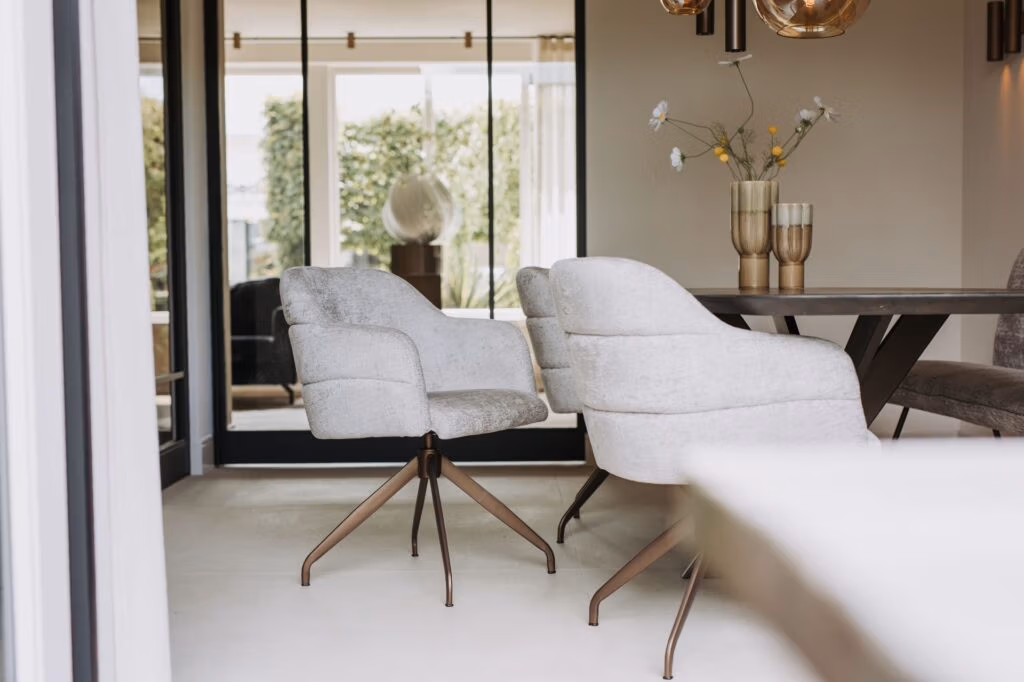 Moderne eethoek met lichtgrijs gestoffeerde stoelen rond een donkerhouten tafel, glazen vazen met bloemen als middelpunt en grote ramen die het groen buiten laten zien. De kamer heeft een minimalistisch, elegant ontwerp.