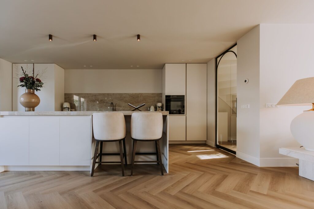 Moderne keuken met witte kasten, een marmeren eiland en twee beige barkrukken, minimalistische inrichting, visgraat houten vloeren, een vaas met bloemen en een grote zwart omlijste glazen deur - perfect voor een minimalistisch interieur.