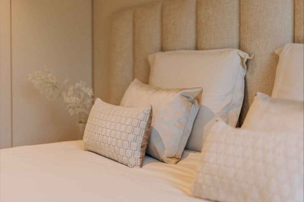 Een netjes opgemaakt bed met een beige gestoffeerd hoofdeinde, versierd met vier crèmekleurige kussens in verschillende maten en texturen. Zachte verlichting versterkt het minimalistische interieur en geeft de kamer een warme en uitnodigende uitstraling.
