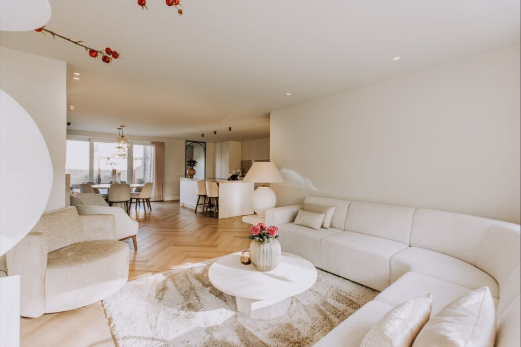 Een lichte, moderne woonkamer met een gebogen witte bank, ronde salontafel met bloemen, neutrale fauteuils en een open keuken en eethoek op de achtergrond, allemaal ingericht in lichte, natuurlijke tinten.