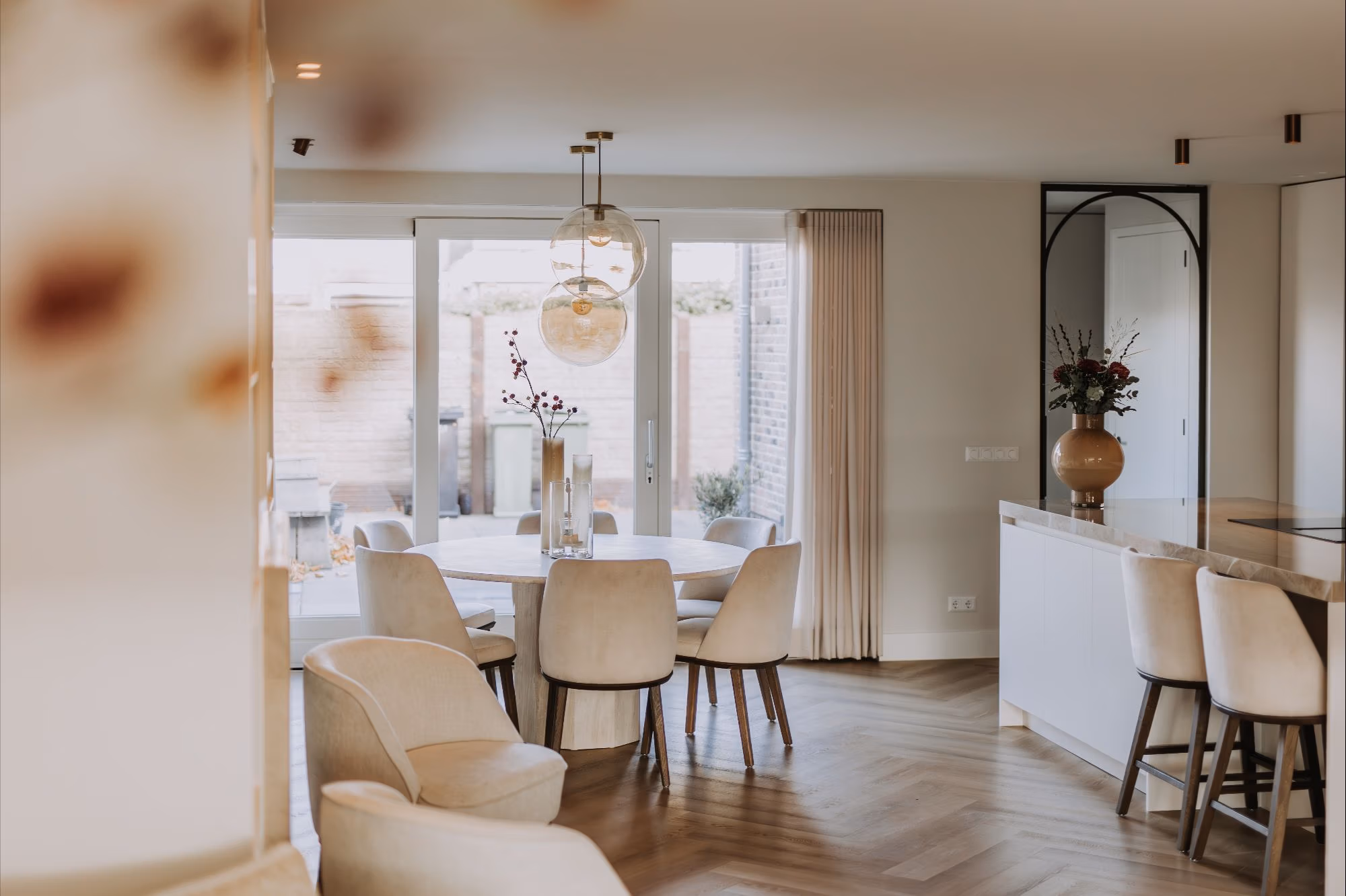 Een moderne eethoek met een ronde tafel en zes crèmekleurige stoelen, hanglampen erboven en grote glazen deuren die leiden naar een patio. De ruimte heeft een minimalistisch ontwerp met neutrale tinten en houten vloeren.