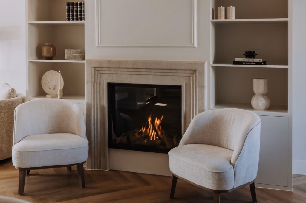 Twee moderne crèmekleurige fauteuils staan tegenover elkaar voor een verlichte open haard, omringd door ingebouwde witte planken versierd met boeken, vazen en minimalistische voorwerpen in een gezellige woonkamer.