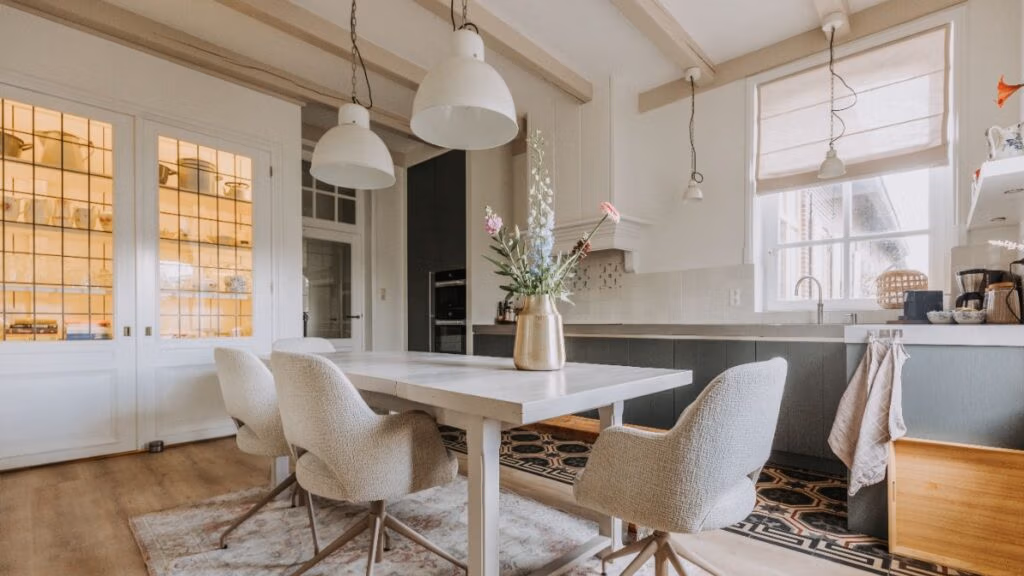 Moderne keuken met een houten tafel, vier stoelen met kussens, een vaas met bloemen, grote hanglampen en natuurlijk licht afkomstig van ramen met beige jaloezieën. Aan de rechterkant zijn planken en apparaten zichtbaar.