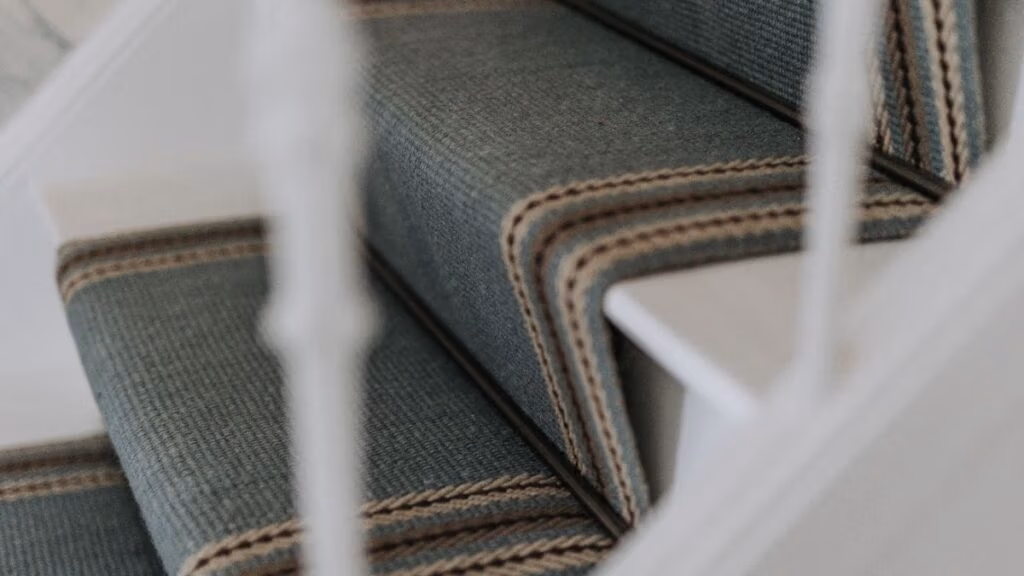 Close-up van een tapijttrap met een blauwgroene loper, afgeboord met beige en bruine strepen en witte trapleuningen die gedeeltelijk zichtbaar zijn op de voorgrond.