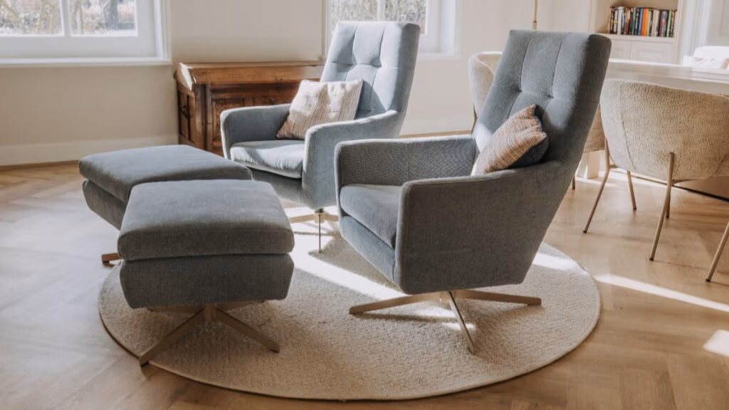 Twee moderne blauwe fauteuils met bijpassende voetenbankjes staan op een rond vloerkleed in een lichte kamer met houten vloeren, grote ramen, een houten kast en een kleine boekenplank op de achtergrond.