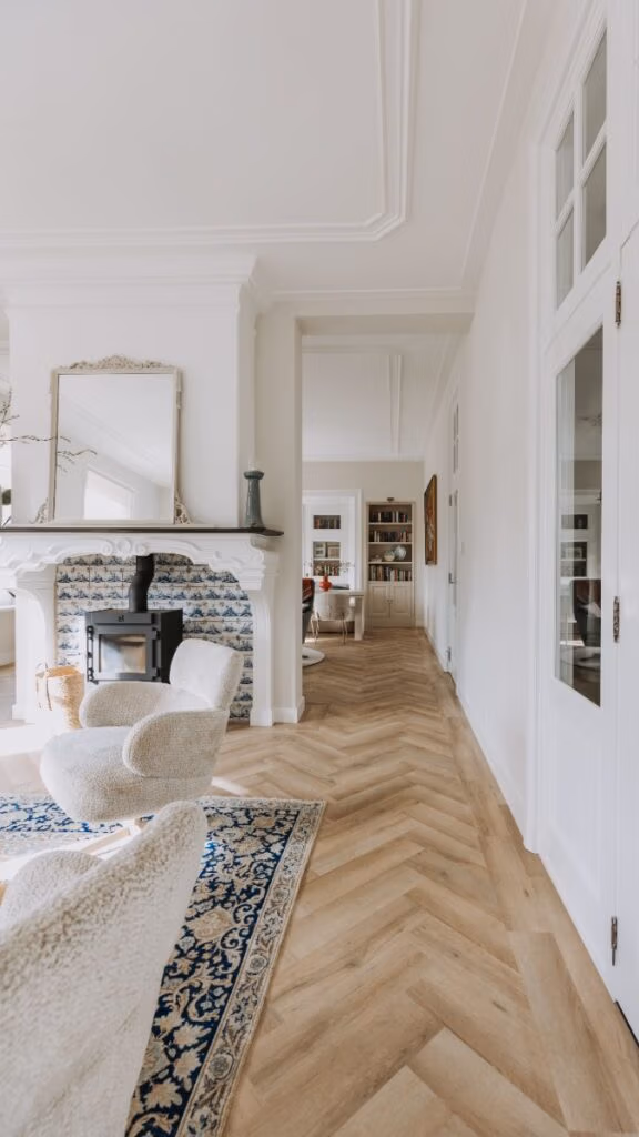 Lichte, elegante woonkamer met chevron houten vloer, twee crèmekleurige fauteuils op een blauw tapijt met patroon, een decoratieve open haard met een grote spiegel en uitzicht op een met boekenplanken beklede kamer daarachter.