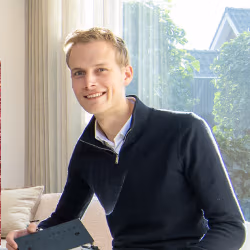 Een jonge man met kort blond haar, draagt een donkere trui over een overhemd met kraag, zit binnenshuis op een bank en glimlacht naar de camera met natuurlijk licht dat door een raam achter hem naar binnen valt.