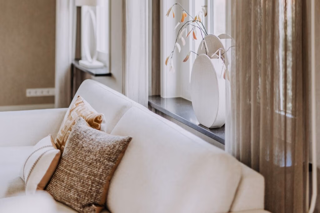 Een moderne woonkamer met een lichtbeige bank, gedessineerde kussens, transparante gordijnen en decoratieve vazen met takken op een vensterbank, die een gezellige en elegante sfeer creëren met zacht natuurlijk licht.