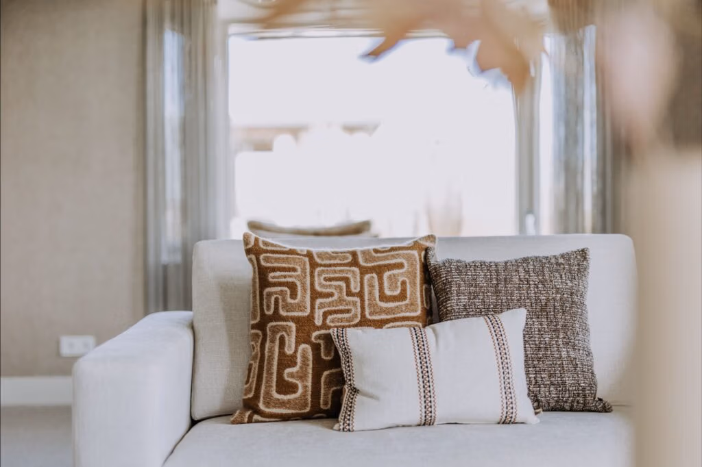 Een lichtgekleurde sofa met drie sierkussens - één met een bruin geometrisch patroon, één met bruine textuur en één wit met bruine strepen - staat in een zacht verlichte, moderne woonkamer met neutrale tinten.