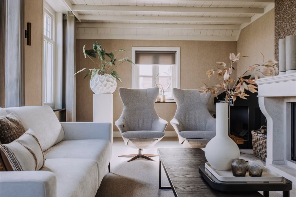 Een moderne woonkamer met neutrale tinten, met een lichte bank, twee grijze draaistoelen, een donkere salontafel met decoratieve vazen, planten en grote ramen die natuurlijk licht binnenlaten.