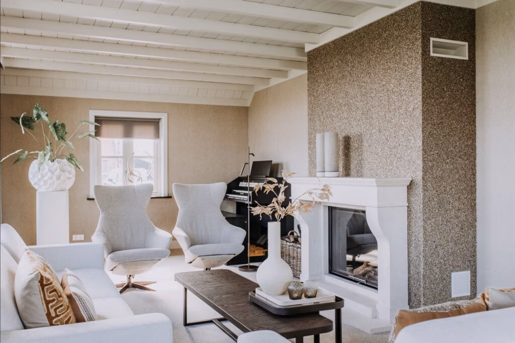 Moderne woonkamer met neutrale tinten, twee grijze fauteuils, een witte bank, een salontafel van donker hout, open haard, decoratieve planten en een piano in de hoek. Licht stroomt naar binnen door een klein raam.
