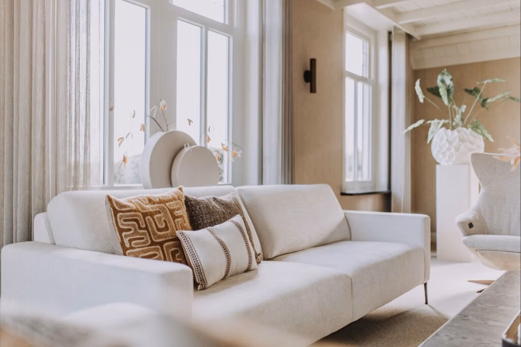Een lichte woonkamer met een lichtbeige bank, kussens met patroon, grote ramen met transparante gordijnen en decoratieve planten op de achtergrond, die een gezellige en moderne sfeer creëren.