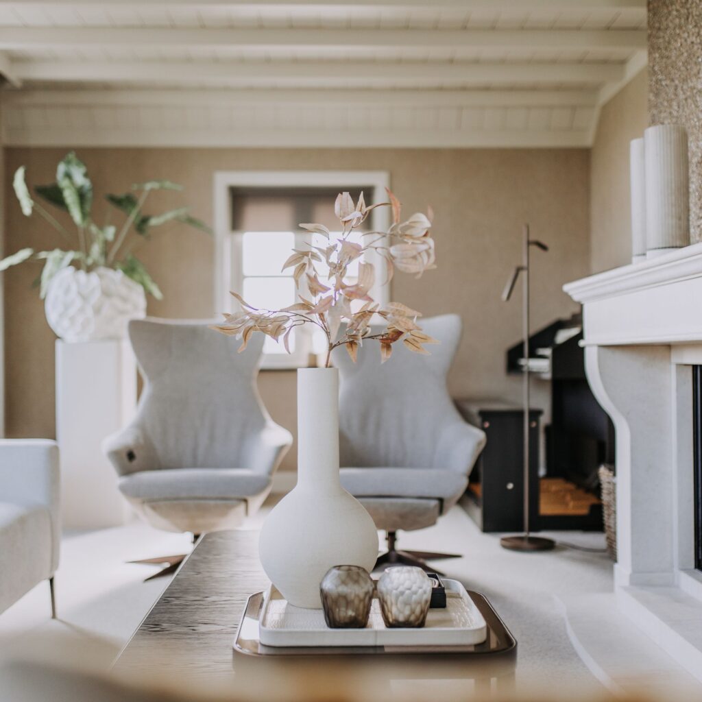 Een moderne woonkamer met lichtgrijze fauteuils, een witte vaas met decoratieve takken op een dienblad en een grote potplant op de achtergrond. De ruimte is licht met natuurlijk licht en neutrale tinten.