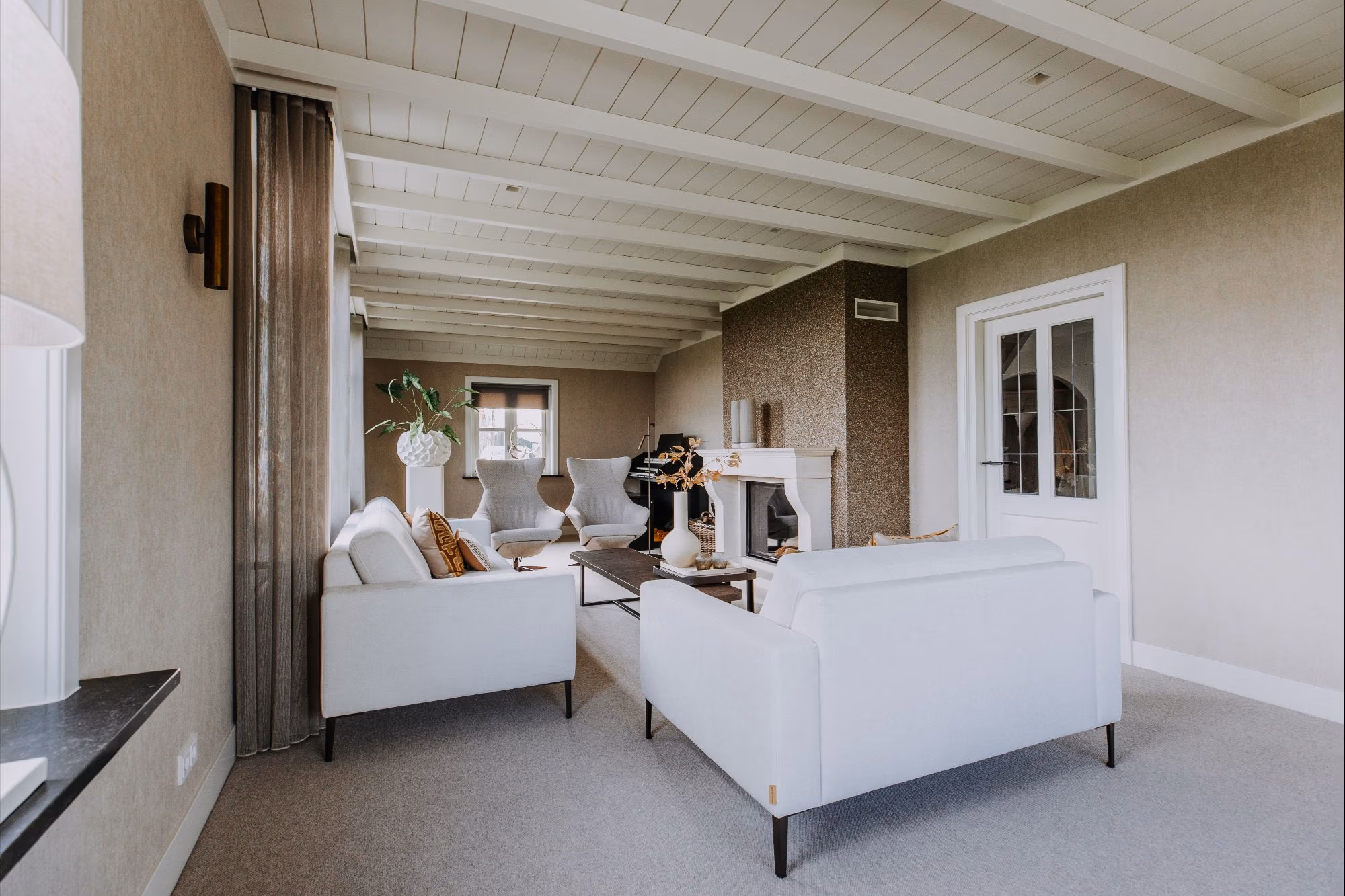 Een lichte, moderne woonkamer met witte banken, een salontafel en een open haard. Neutrale tinten domineren de ruimte, met grote ramen en zichtbare plafondbalken die bijdragen aan de luchtige, uitnodigende sfeer.
