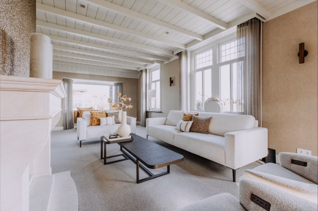 Een moderne, lichte woonkamer met witte banken, een zwarte salontafel, grote ramen, een neutrale inrichting en lichtbeige muren en plafondbalken. Twee fauteuils en decoratieve vazen maken de gezellige, elegante ruimte compleet.