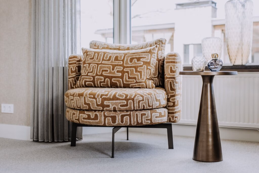 Een moderne fauteuil met een gedurfd geometrisch patroon in beige en wit staat naast een ronde bijzettafel met glazen vazen, in een lichte kamer met grote ramen en lichtgrijze gordijnen.