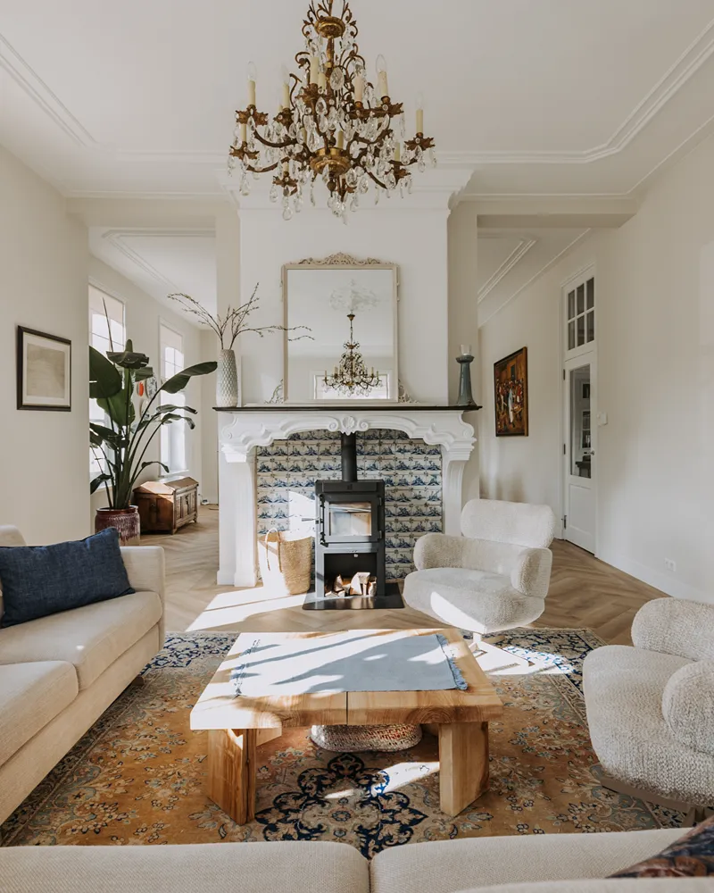 Elegante woonkamer met neutrale fauteuils, een houten salontafel, een sierlijk tapijt, een kroonluchter, een open haard met decoratieve tegels, een grote spiegel en een hoge groene plant bij het raam. Het zonlicht stroomt naar binnen en zorgt voor een warme, uitnodigende sfeer die de balans in je interieur perfect weerspiegelt.