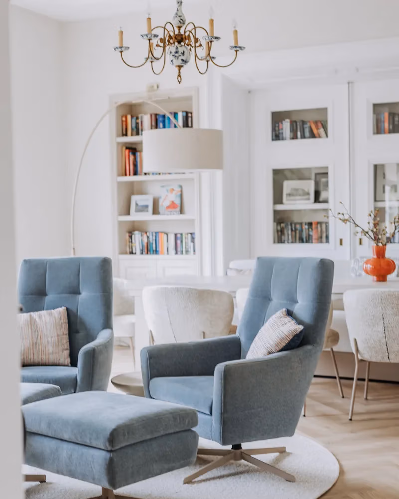 Moderne woonkamer met twee blauwe fauteuils, een bijpassende poef, een kroonluchter, boekenkasten met boeken, een ronde witte tafel, witte stoelen en een vaas met bloemen op de tafel. De lichte en luchtige inrichting in combinatie met de houten vloer zorgt voor een perfecte balans in je interieur.