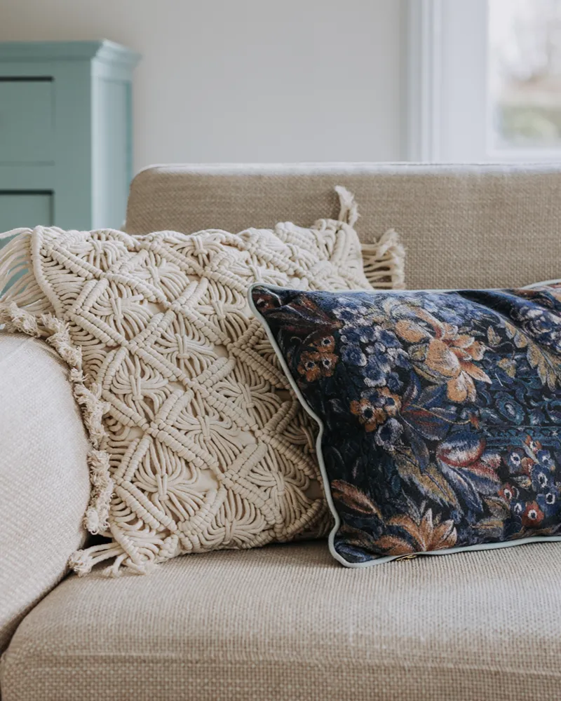 Twee sierkussens op een beige bank zorgen voor evenwicht in je interieur: het ene met een crèmekleurig macramé-patroon en franjes, het andere met een donker bloemenpatroon in tapijtstijl. Op de achtergrond is een deel van een lichtblauwe kast te zien.