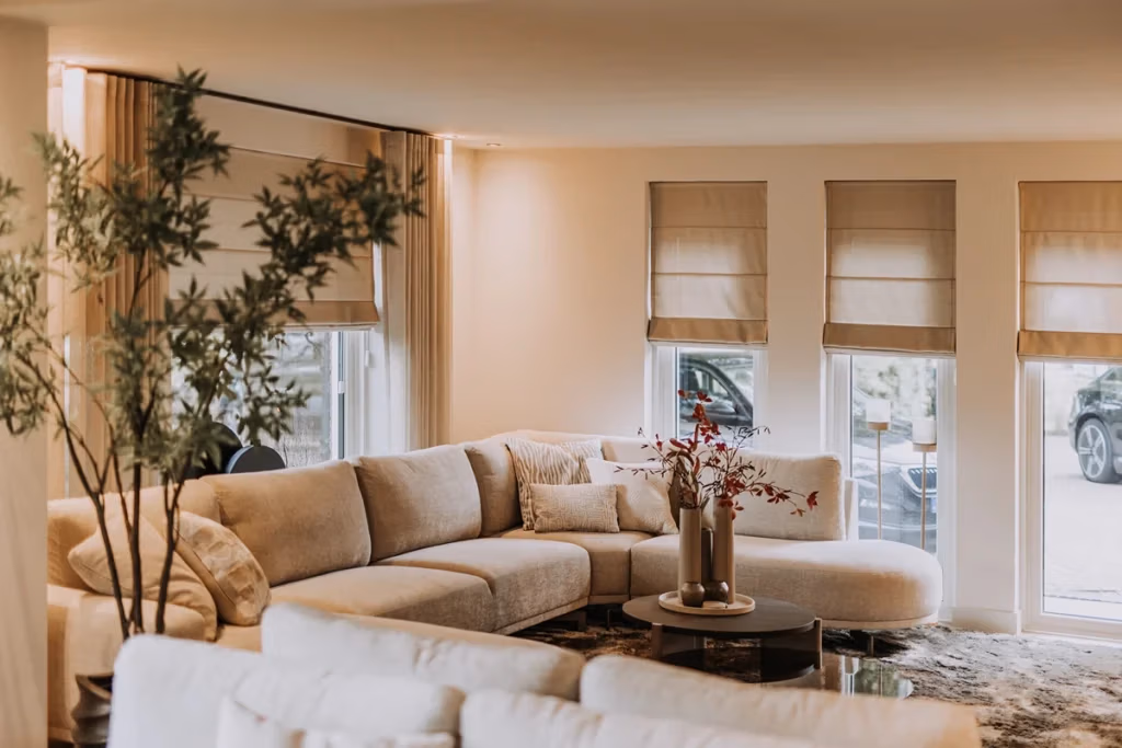 Moderne woonkamer met een grote beige hoekbank, een ronde salontafel met een vaas met bloemen, een hoge kamerplant en drie ramen met beige vouwgordijnen. Natuurlijk licht vult de gezellige ruimte in neutrale tinten en biedt volop woonkamerinspiratie voor wie op zoek is naar een ‘Hotel Chic’-look.