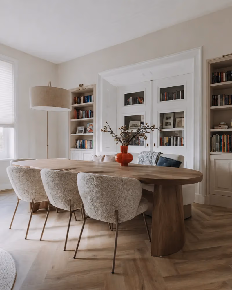 Een moderne eetkamer met een ovale houten tafel, vier stoelen met een structuur, een rode vaas met takken, een staande lamp, ingebouwde boekenkasten en dubbele deuren in een lichte, neutrale ruimte zorgt voor een perfecte balans in je interieur.
